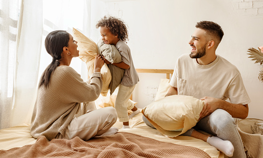 happy family multiethnic mother, father and son  laughing, playing, fights pillows and jumping in bed in bedroom at home