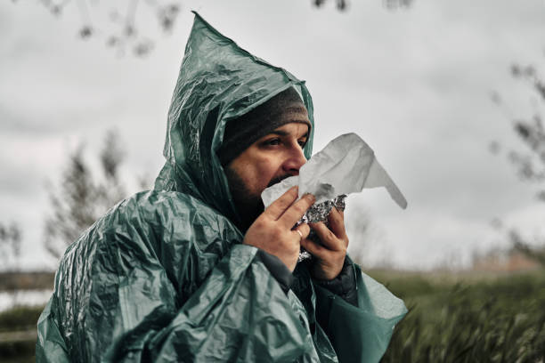 um homem com barba em uma capa de chuva verde come na natureza. tempo nublado, fast food. - 11313 - fotografias e filmes do acervo