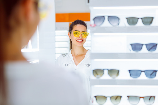 Reflection of young woman in eyewear retail trying on yellow sunglasses and smiling