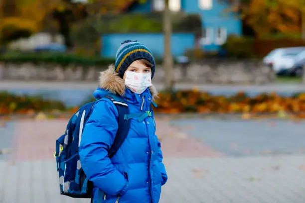 Little kid boy wearing medical mask on the way to school. Child backpack satchel. Schoolkid on cold autumn or winter day with warm clothes. Lockdown and quarantine time during corona pandemic disease.