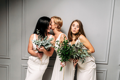 Boho style wedding. Bridesmaids in long ivory dresses, in their hands green bouquets. They are whispering among themselves. Photo in studio on a gray background
