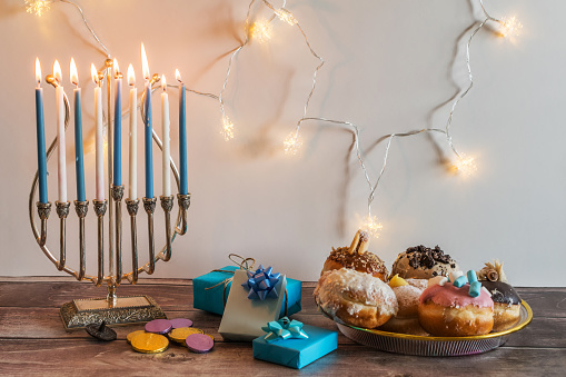 Concept of of jewish religious holiday hanukkah with Menorah (Judaism candelabra), gifts, sufganiyot (doughnuts), spinning top toys (dreidel), and chocolate coins, on wooden table. Front view.