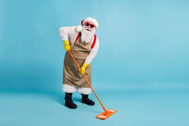 Full body size photo of retired old man grey beard hold mop washing, floor public janitor wear santa x-mas costume apron rubber glove suspender sunglass cap isolated blue color background