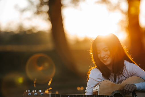 One carefree beautiful young woman with long hair playing acoustic guitar and enjoying picnic day in nature at beautiful sunset in background