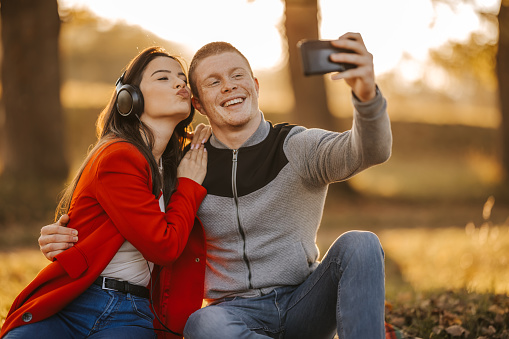 Cheerful young heterosexual couple sitting and taking selfie on mobile phone on picnic blanket in nature at sunset and having fun while listening good music on headphones