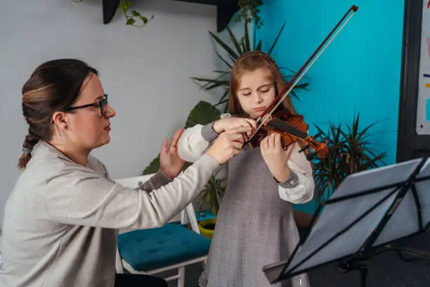 Little girl is frowning while female teacher showing how to play violin in the classroom.