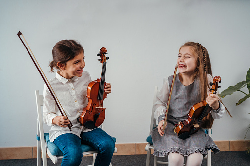 Little girls are sitting while holding their violins and laughing in the classroom.