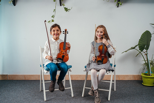 Smiling little girls are sitting while holding their violins and looking at camera in the classroom.