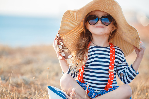 Happy little girl with long blond hair in a straw hat and sunglasses walks on a meadow at sunset with a seascape on the background.A funny child plays alone in nature while sitting on dry grass near a pond.Happy childhood concept, out of size things