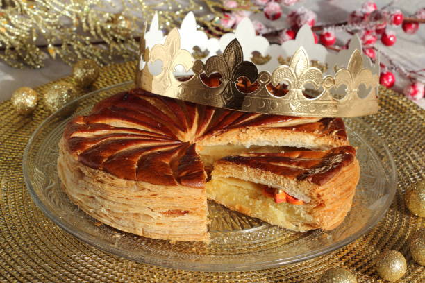 Epiphany kings cake with a golden paper crown and one small porcelain good look charm inside. Galette des rois. Epiphany cake with a golden paper crown and one small porcelain  charm inside on a glass plate on the holiday table. Slice of kings cake. Galette des rois. galette stock pictures, royalty-free photos & images