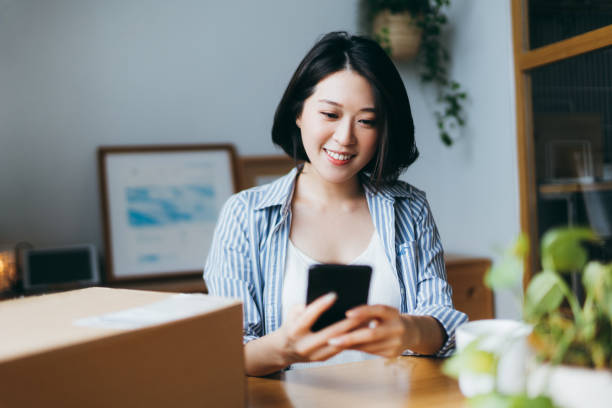 istock Beautiful smiling young Asian woman preparing a parcel for delivery using mobile app device on smartphone at home. Technology makes life so much easier 1284021209