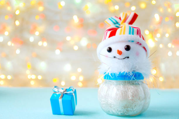 istock Toy funny snowman in a colored hat and a blue gift box with a ribbon on a light background with bokeh. Background for greeting cards, happy new year and Christmas, space for text 1284021307