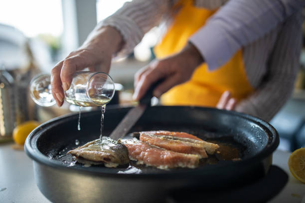 senior woman adding white wine while preparing fish with her husband at home - cooking senior adult healthy lifestyle couple imagens e fotografias de stock