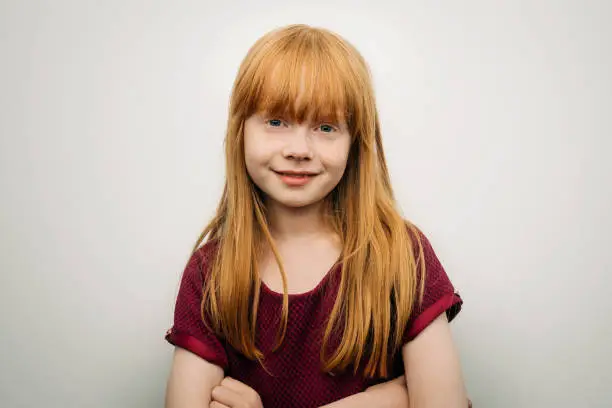 Portrait of cute smiling girl with redhead. Female child is with arms crossed. She is against white background.