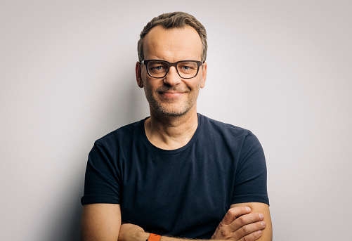 Portrait of confident mature man in t-shirt. Smiling handsome male is wearing eyeglasses. He is with arms crossed against white background.