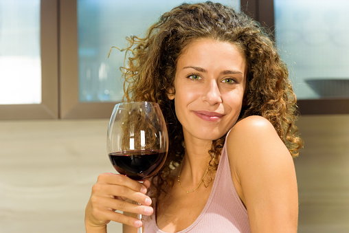 Attractive friendly young woman with a lovely warm smile toasting the camera with a large elegant glass of red wine to celebrate in a close up portrait