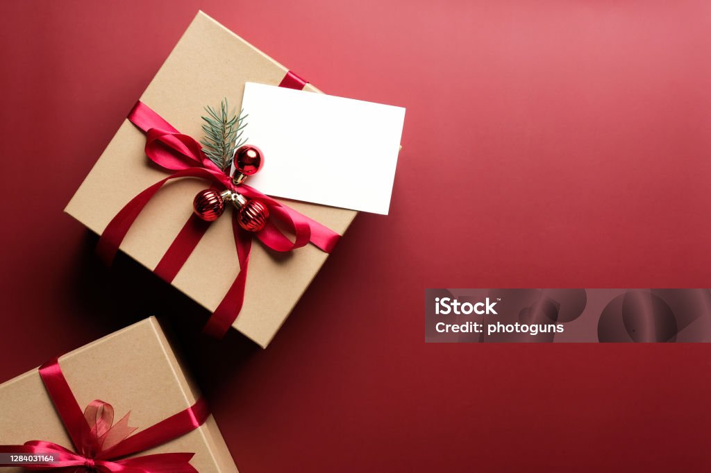 Carton gift box decorated red ribbon bow and blank paper card mockup on marsala red background. Flat lay, top view. Christmas gift, New Year present concept. Christmas Present Stock Photo