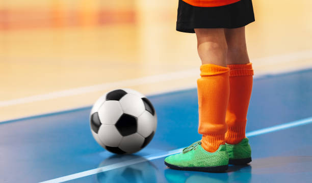 giocatore di calcio indoor con palla classica. formazione sul futsal per bambini. gambe di giovane giocatore di futsal in tacchette da calcio e calzini. palazzetto dello sport al coperto. giocatore in uniforme arancione. background sportivo di livello juni - futsal indoors soccer ball soccer foto e immagini stock