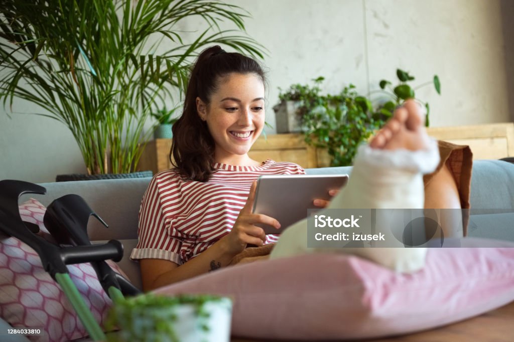Cheerful young woman with broken leg at home Smiling young man with broken leg in plaster cast lying down on sofa at home and using a digital tablet. Orthopedic Cast Stock Photo