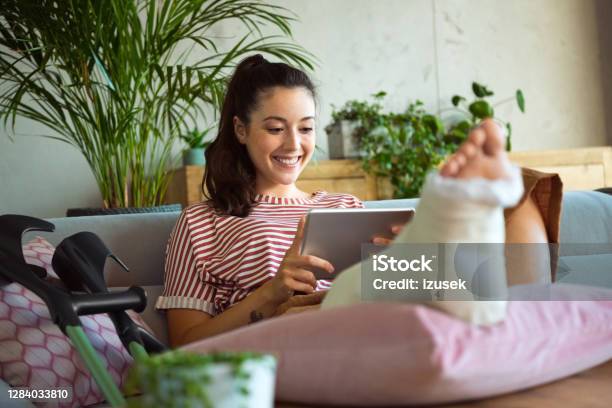 Cheerful Young Woman With Broken Leg At Home Stock Photo - Download Image Now - Orthopedic Cast, Broken Leg, Adult