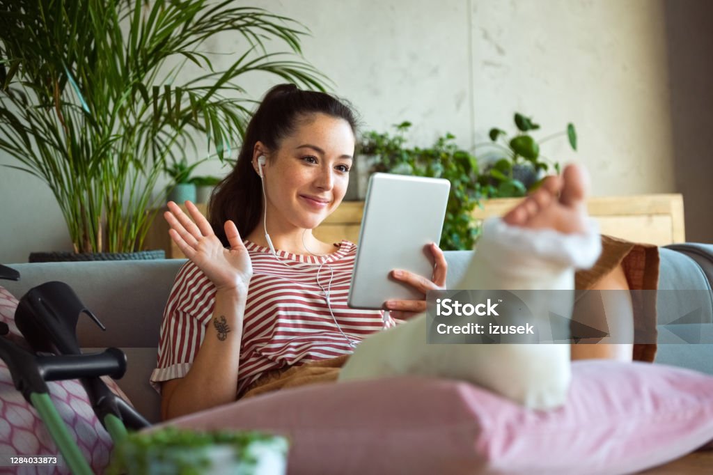 Cheerful young woman with broken leg during video call Smiling young man with broken leg in plaster cast lying down on sofa at home, using a digital tablet and having video conference. Broken Leg Stock Photo