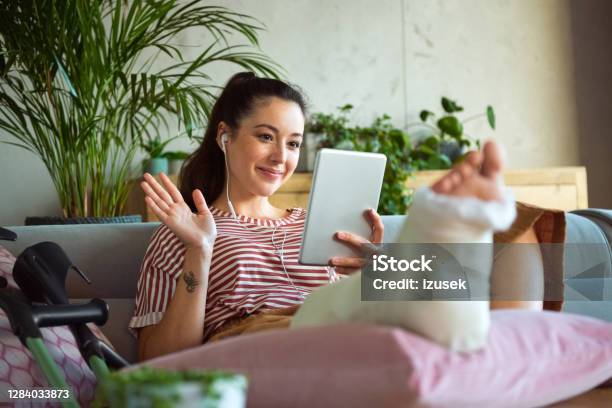 Cheerful Young Woman With Broken Leg During Video Call Stock Photo - Download Image Now