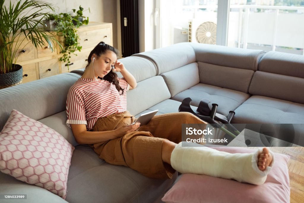 Sad young woman with broken leg at home Worried young man with broken leg in plaster cast lying down on sofa at home and using a digital tablet. Broken Leg Stock Photo