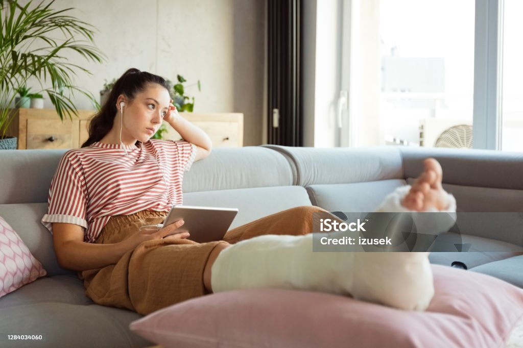 Sad young woman with broken leg at home Worried young man with broken leg in plaster cast lying down on sofa at home, holding a digital tablet and looking through window. Broken Leg Stock Photo