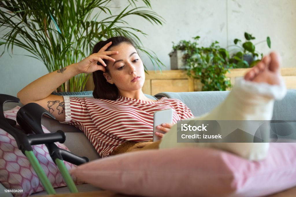 Sad young woman with broken leg using smart phone Worried young man with broken leg in plaster cast lying down on sofa at home and using a smart phone. Broken Leg Stock Photo
