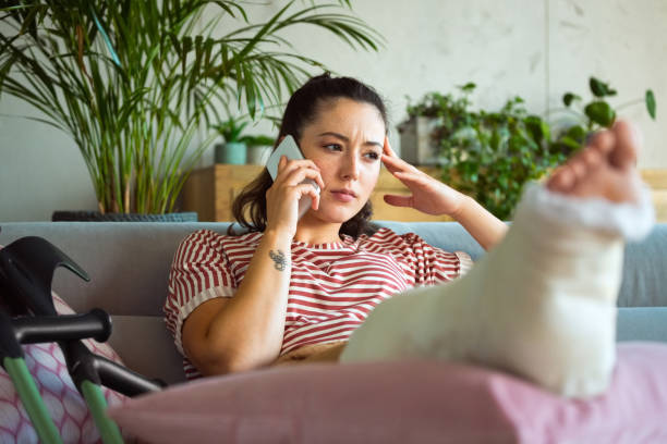 jeune femme inquiète avec la jambe cassée utilisant le téléphone intelligent - insurance and accident themes photos et images de collection