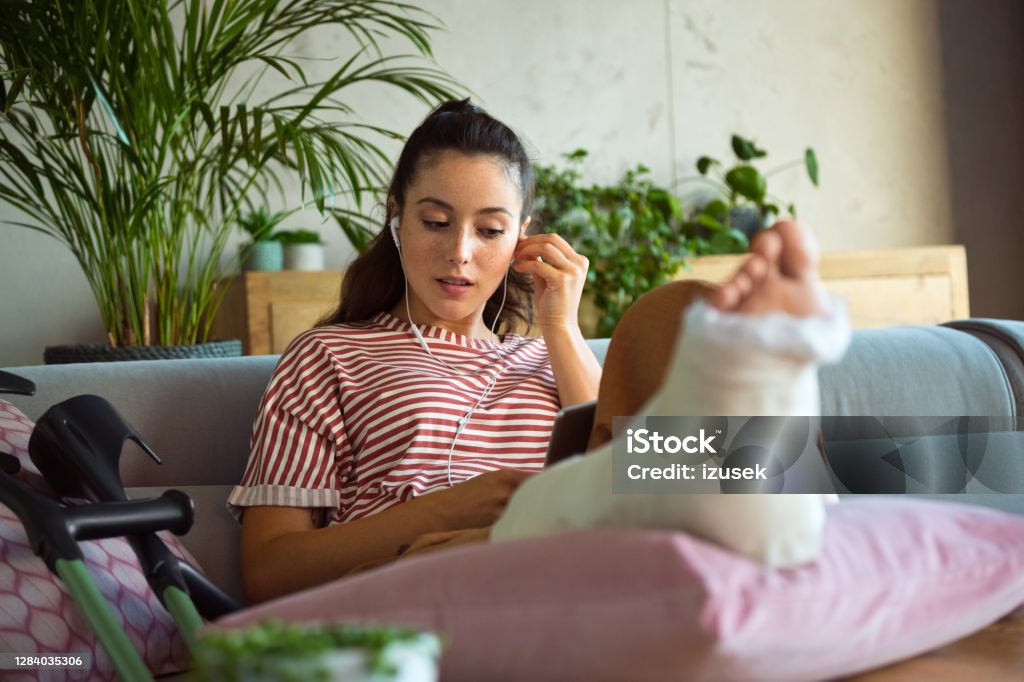 Young woman with broken leg during video call Young man with broken leg in plaster cast lying down on sofa at home, using a digital tablet and having video conference. Recovery Stock Photo