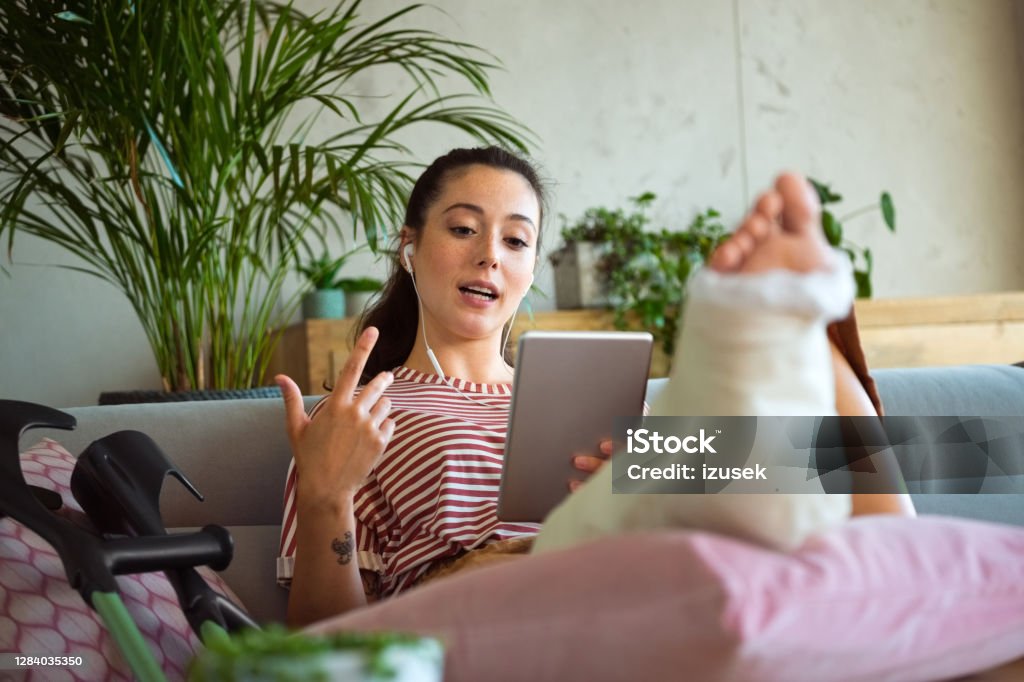 Young woman with broken leg during video call Smiling young man with broken leg in plaster cast lying down on sofa at home, using a digital tablet and having video conference. Telemedicine Stock Photo