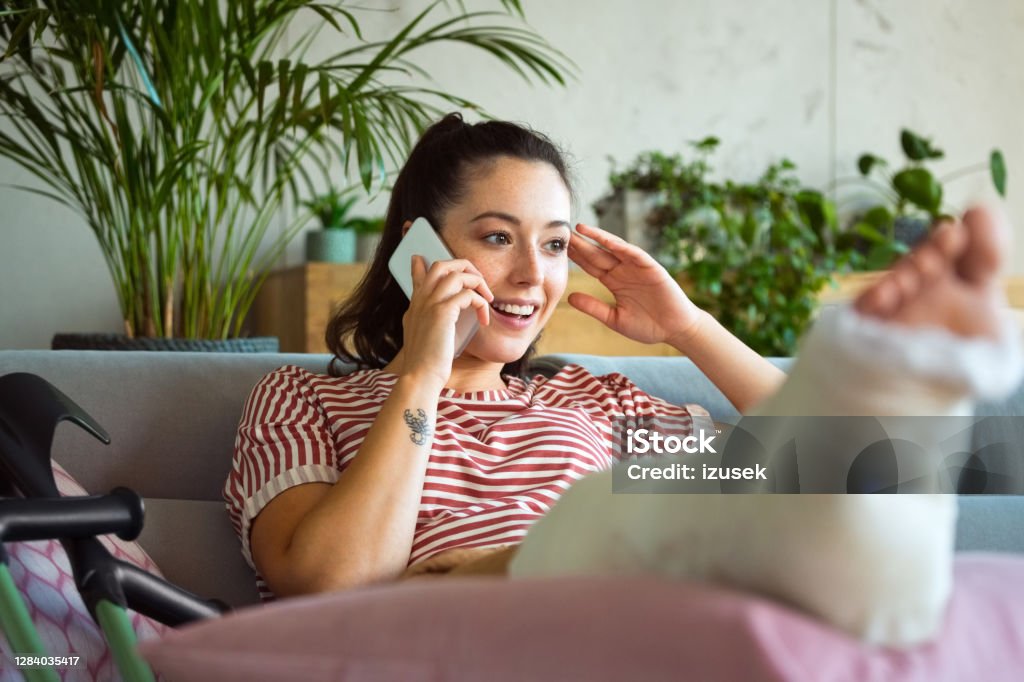 Young woman with broken leg talking on phone Cheerful young man with broken leg in plaster cast lying down on sofa at home and using a smart phone. Broken Leg Stock Photo