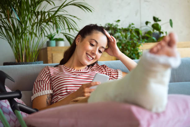 Young woman with broken leg using smart phone Cheerful young man with broken leg in plaster cast lying down on sofa at home and using a smart phone. orthopedic cast stock pictures, royalty-free photos & images