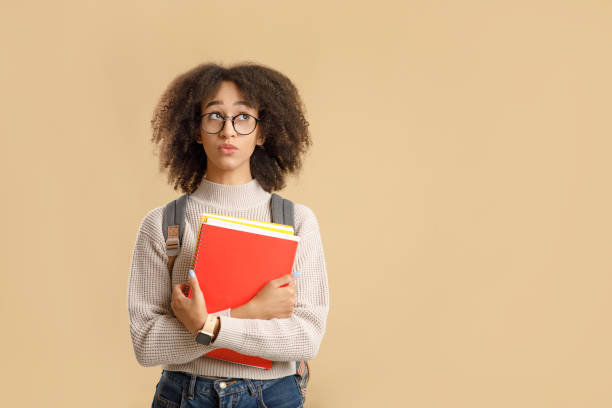 istock Make choices or think about study, problems at education. Pensive african american woman student in glasses with backpack holds notepads and looks up to free space 1284037575