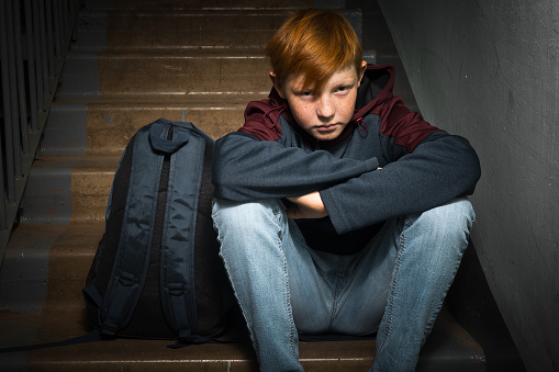 A red-haired boy, a teenager of twelve, with an anxious expression on his face, sits on the ladders at the entrance of a residential building. Children's fear and anxiety, family social problems.