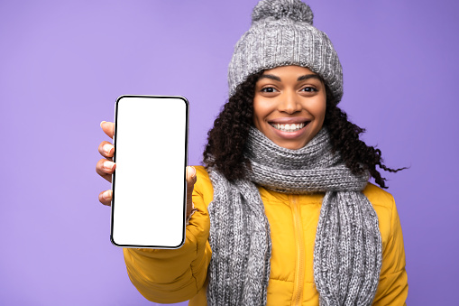 African Woman Showing Smartphone Empty Screen Recommending Mobile Phone Application Posing Wearing Winter Jacket Standing Over Purple Studio Background. Cellphone Display Mockup Advertisement