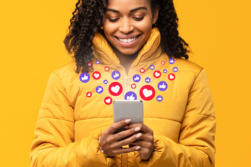 Mujer negra feliz sosteniendo el teléfono inteligente con botones del me gusta, fondo amarillo photo
