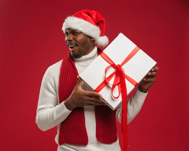 istock A dark-skinned man in a sweater and christmas hat holds a gift box and grins 1284041311