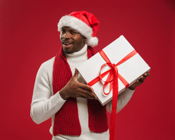 istock A dark-skinned man in a sweater and christmas hat holds a gift box and grins 1284041367