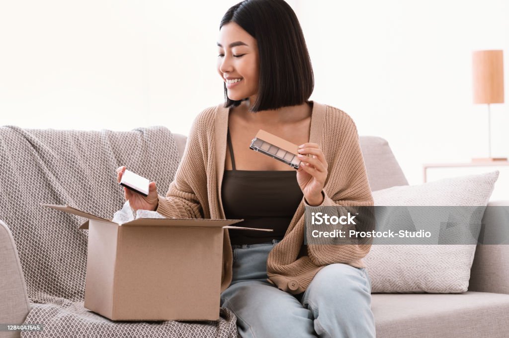 Happy asian lady unpacking parcel with beauty box Beauty Box Concept. Portrait of smiling asian lady received delivery package, holding eyeshadow palette for makeup, unpacking cardboard parcel with cosmetics product, sitting on the couch at home Make-Up Stock Photo