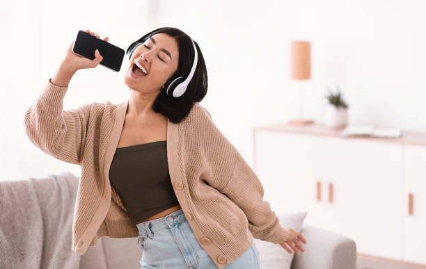 Happy asian lady singing using smartphone as mic Karaoke App. Portrait of emotional asian woman singing her favorite song, dancing and using cellphone as mic, wearing wireless headphones. Excited lady having fun at home in living room, copy space singing stock pictures, royalty-free photos & images