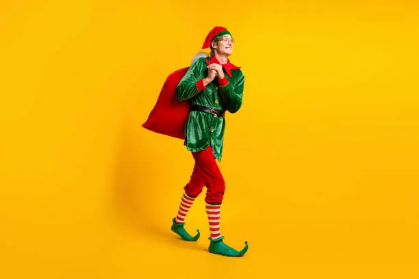 Full length body size view of his he nice attractive cheerful cheery funny guy elf walking carrying sack, wintertime day delivery isolated over bright vivid shine vibrant yellow color background