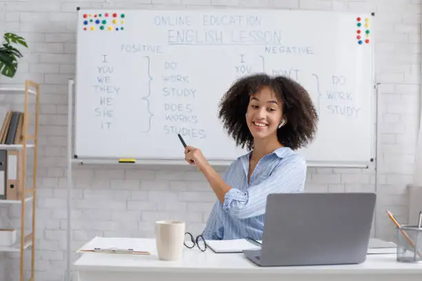 Repetition of rule of English language on online lesson. Smiling african american woman points with marker at whiteboard and watch on laptop webcam sitting at table in classroom interior at home