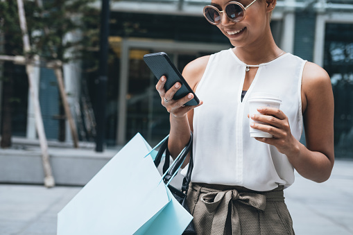 Close up portrait of beautiful smiling short hair blonde woman standing in front of shopping mall. She is typing message on smartphone and holding shopping bag and coffee