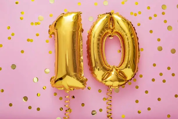 Photo of 10 air balloon numbers on pink background. 10 k gold foil balloons with confetti. Birthday party flat lay with copy space