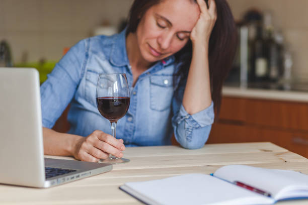 Depressed woman drinking red wine at home. Depressed mid adult woman feeling drunk while drinking wine in the kitchen. Focus is on hand and wineglass. alcohol abuse stock pictures, royalty-free photos & images
