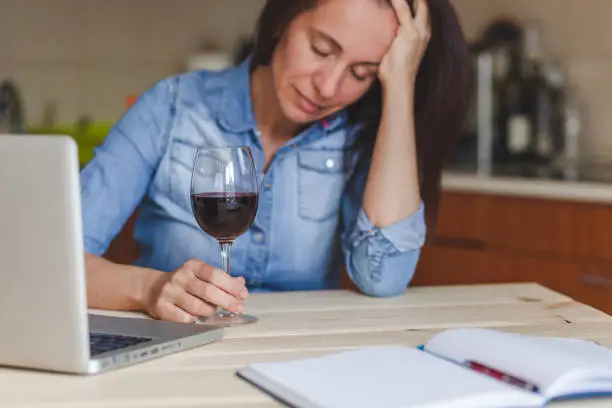 Depressed mid adult woman feeling drunk while drinking wine in the kitchen. Focus is on hand and wineglass.