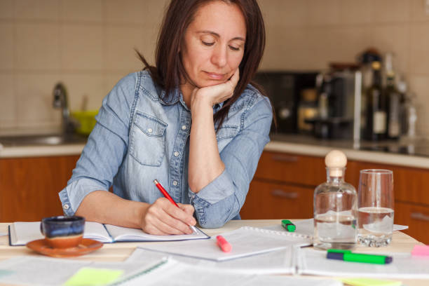 istock Concentrated mid adult woman taking notes while studying from textbook at home. 1284052802