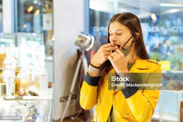 Young And Happy Woman Wearing Protective Face Mask Eating A White Chocolate Standing Inside A Shopgirl Having A Sweet Candy In Autumn After Lockdown Reopeningconcept About Joy And Hapinessnew Normal Lifetstyle Stock Photo - Download Image Now